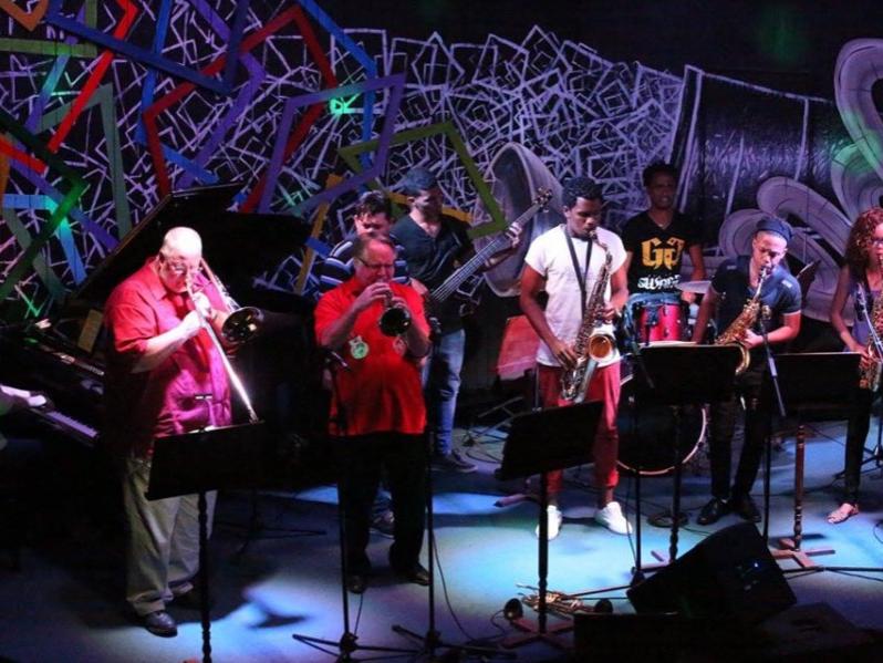 Musicians of varying ages stand on stage in front of microphones and play their instruments.