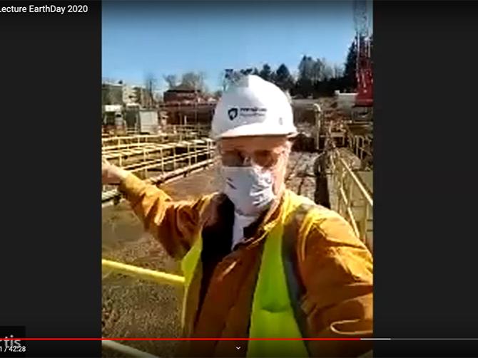 A man wearing a mask, hard hat, and safety glasses gestures to outdoor water pools in a screenshot of a YouTube video.