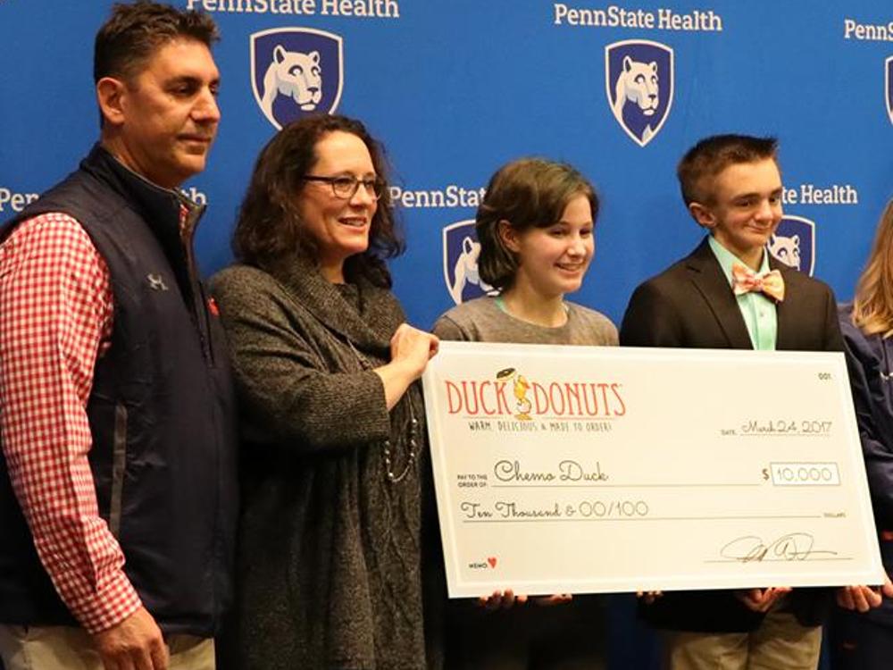Four people pose holding an oversized check. They stand in front of a blue backdrop with the Penn State Health logo.