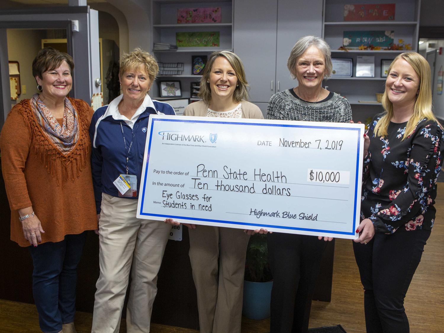 Five women pose with an oversized check in the amount of $10,000, made out from Highmark to Penn State Health. The Memo line reads: Eye glasses for students in need. A desk and office shelves are in the background.
