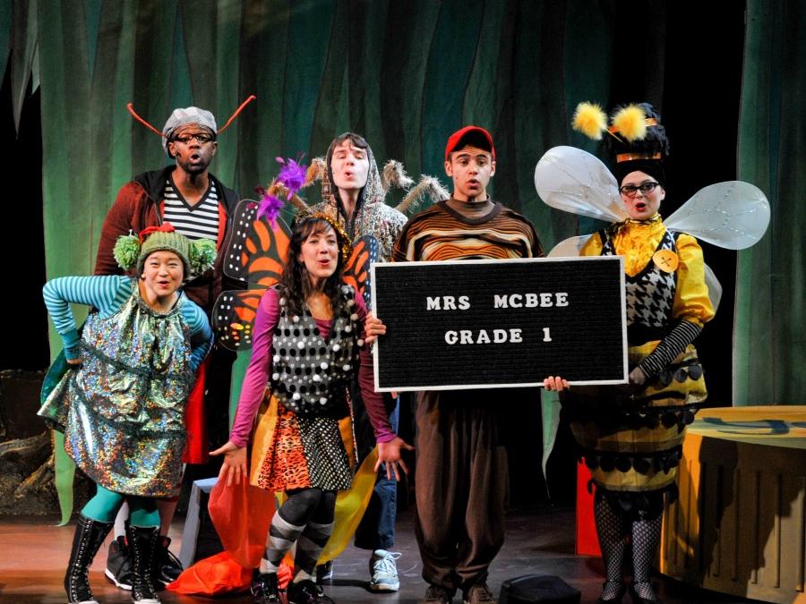 Actors dressed in street clothes to resemble insects stand in a huddle while a character dressed like a worm holds a sign.