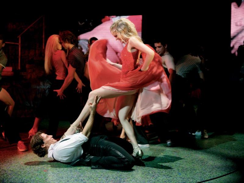 An actress, swishing her dress back and forth, places her left leg atop a male dancer's chest while he lies on his back with his legs bent at the knee and folded underneath him.