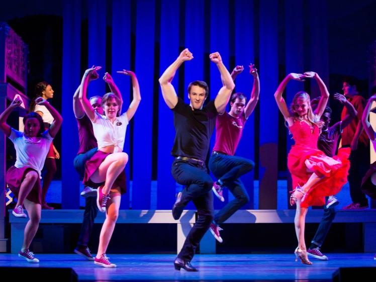A mix of male and female dancers bend their right legs up and extend their arms above them in a dance routine.