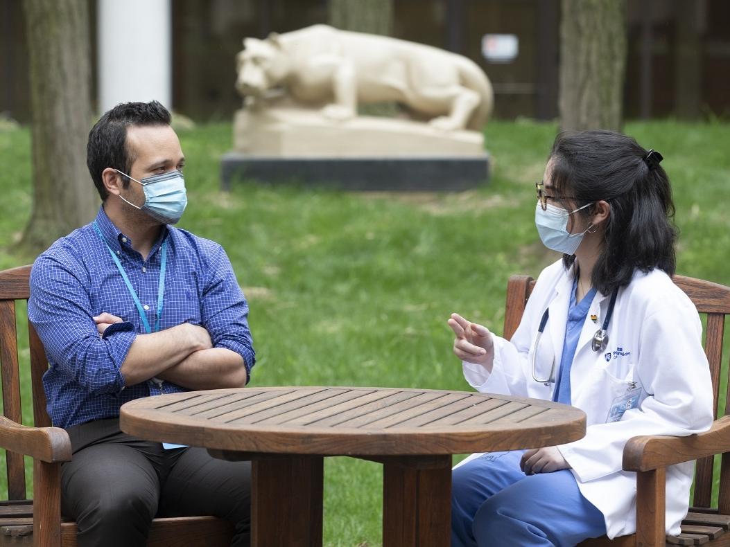 A man and woman sit outside at a wooden table with a statue of the Penn State Nittany Lion in the background. They both wear face masks and the woman is wearing glasses and hospital scrubs under a lab coat. The man’s arms are crossed.