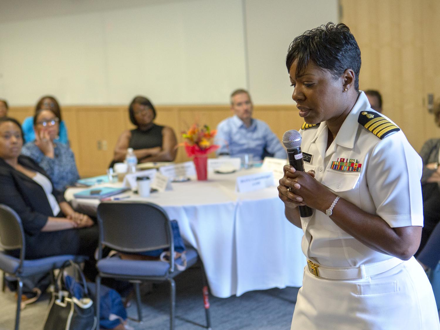 A woman in a military uniform holds a wireless microphone and speaks in front of a group of people.