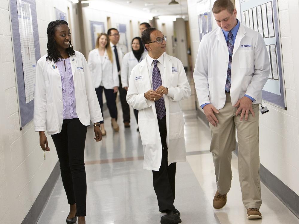 Two men and a woman in white lab coats walk through a tile-floored hallway. Another group of four people are in the background behind them.