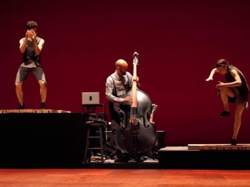 A musician playing an upright bass is flanked by tap dancers on short risers dancing atop electronic trigger boards.