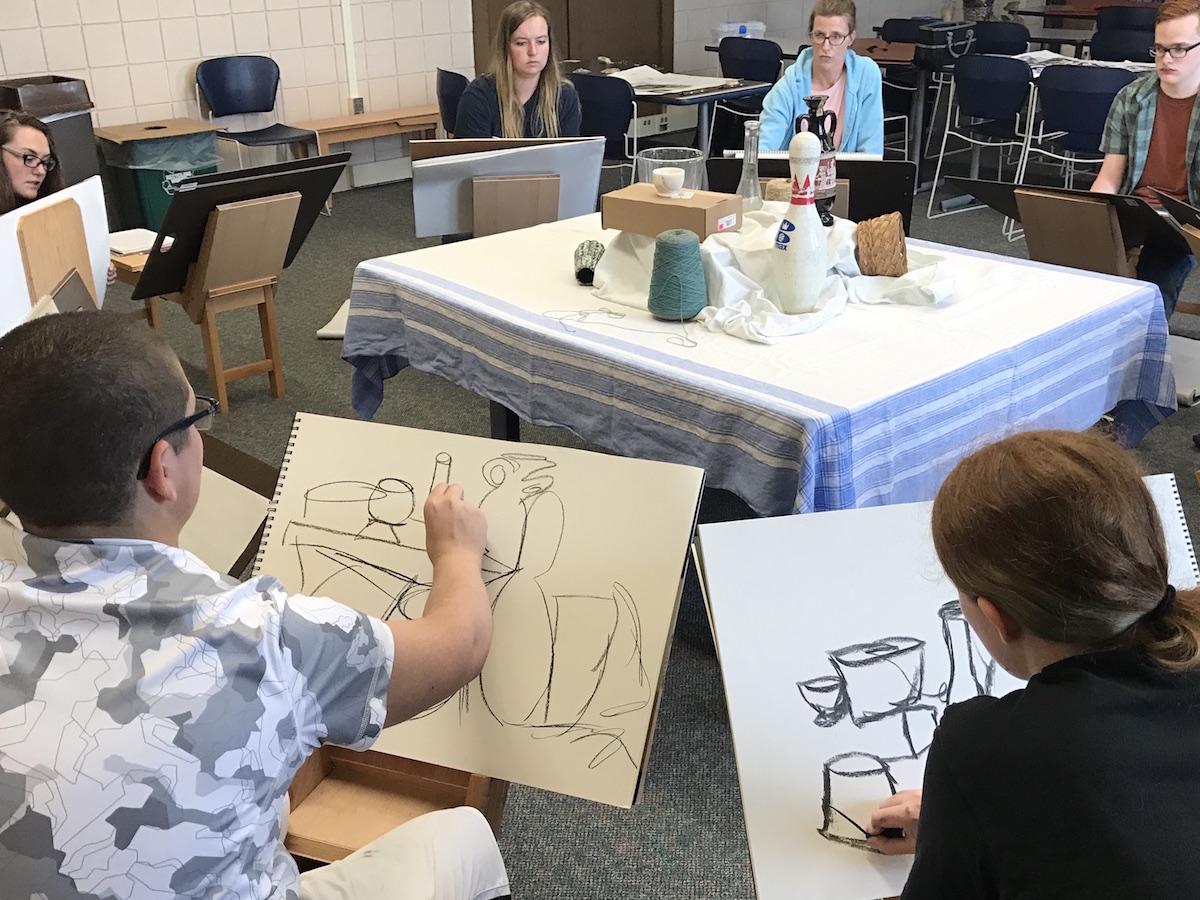 Students sit on drawing horses and sketch a still life with charcoal.