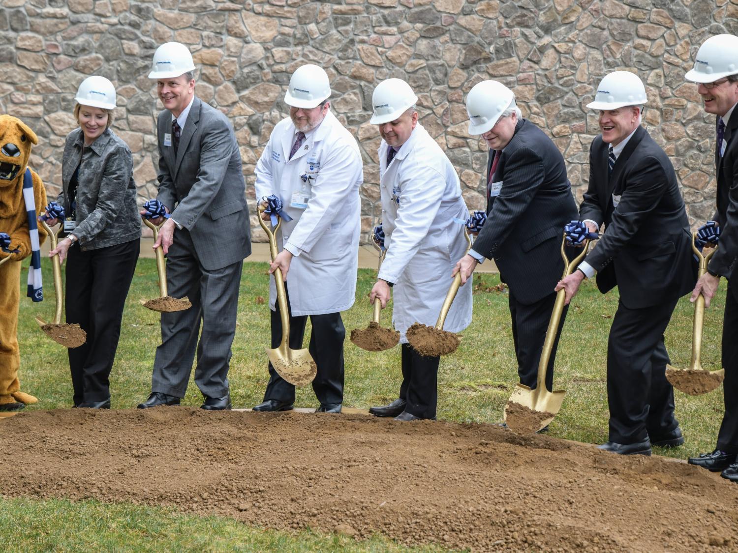 Seven people -- five in business suits, two in white physician's coats, and all wearing white helmets -- use gold shovels to dig into a pile of dirt. A stone wall is in the background.