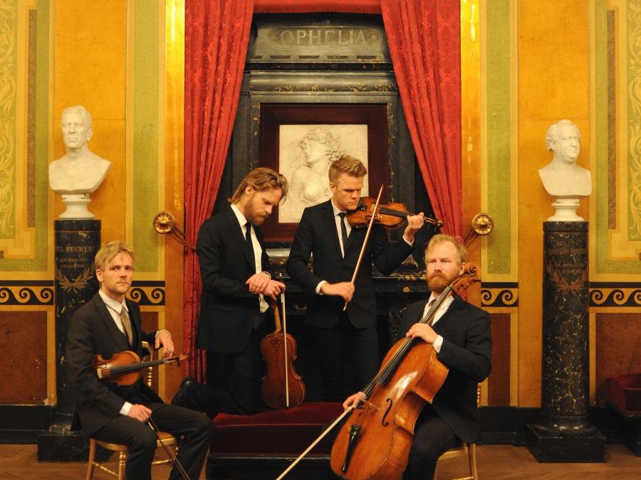 Four chamber musicians sit or kneel in from of a curtain and marble artworks while they play their stringed instruments.