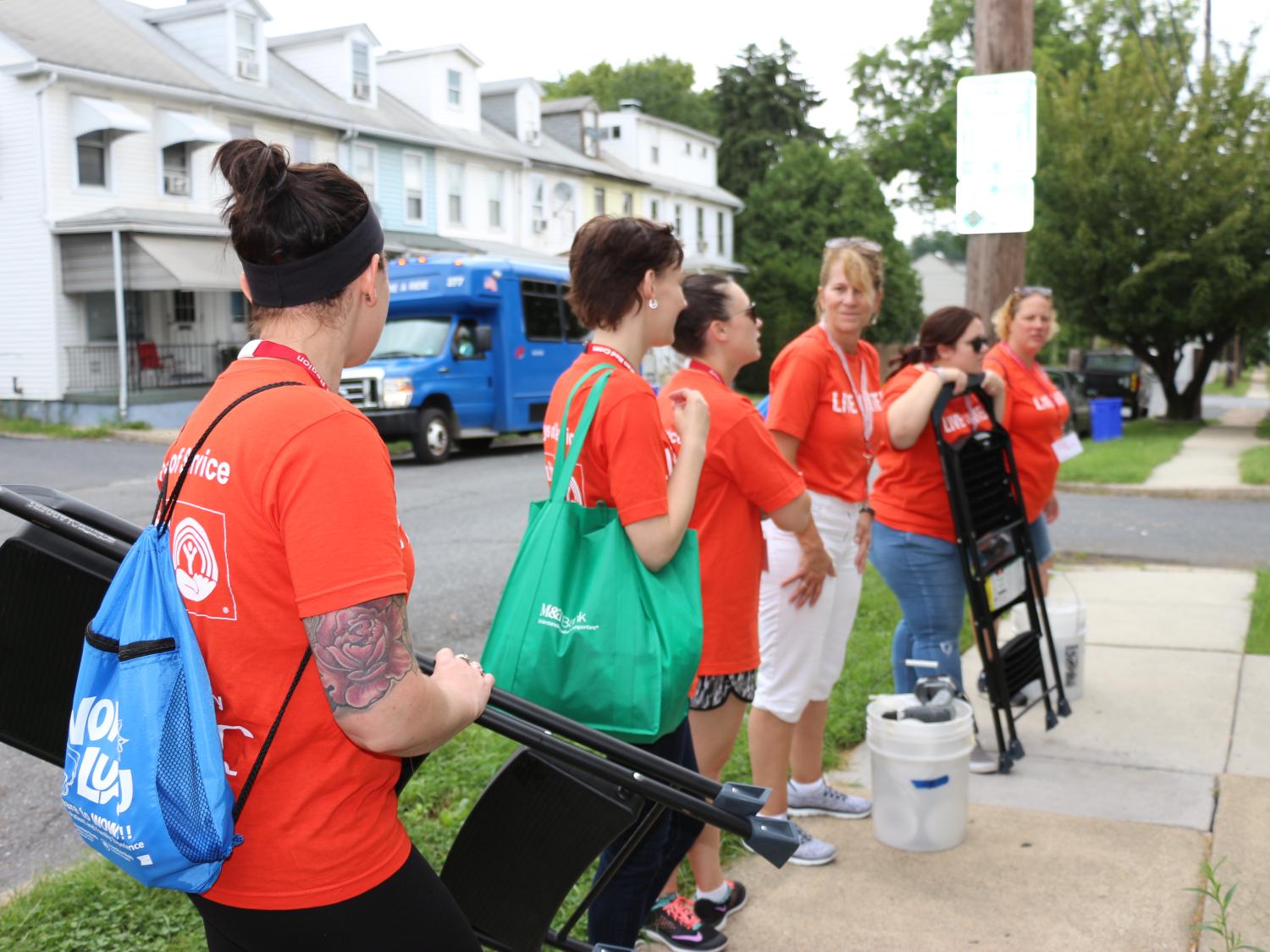 Six women stand along a sidewalk near a city street. Some hold bags; two are holding step ladders. They all wear orange shirts with the slogan “LIVE UNITED” and the United Way logo.