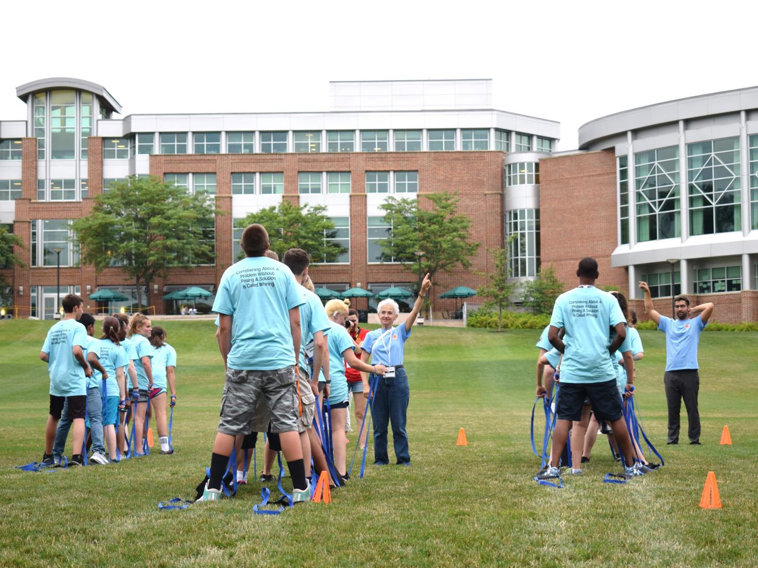 Students participate in a Recreation/Team Building centipede challenge, attempting to walk together on two cloth planks with straps for handles.