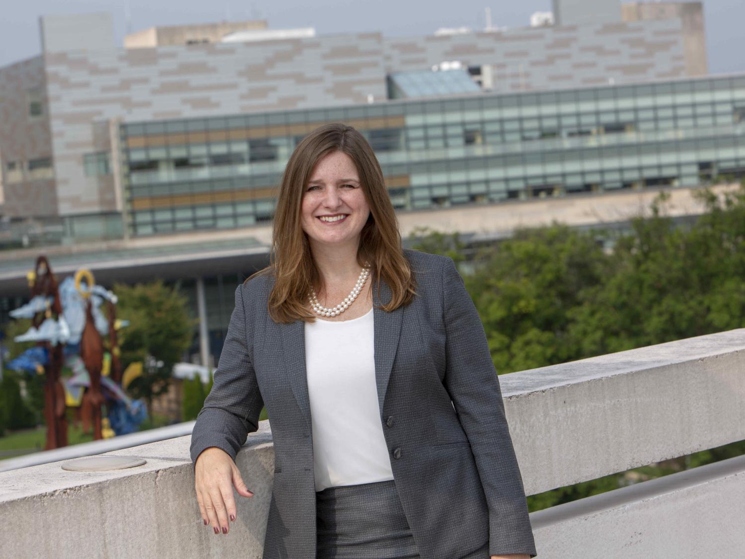 Deborah Berini poses for a photo wearing professional attire. She leans on a concrete wall, and Hershey Medical enter is in the near background.