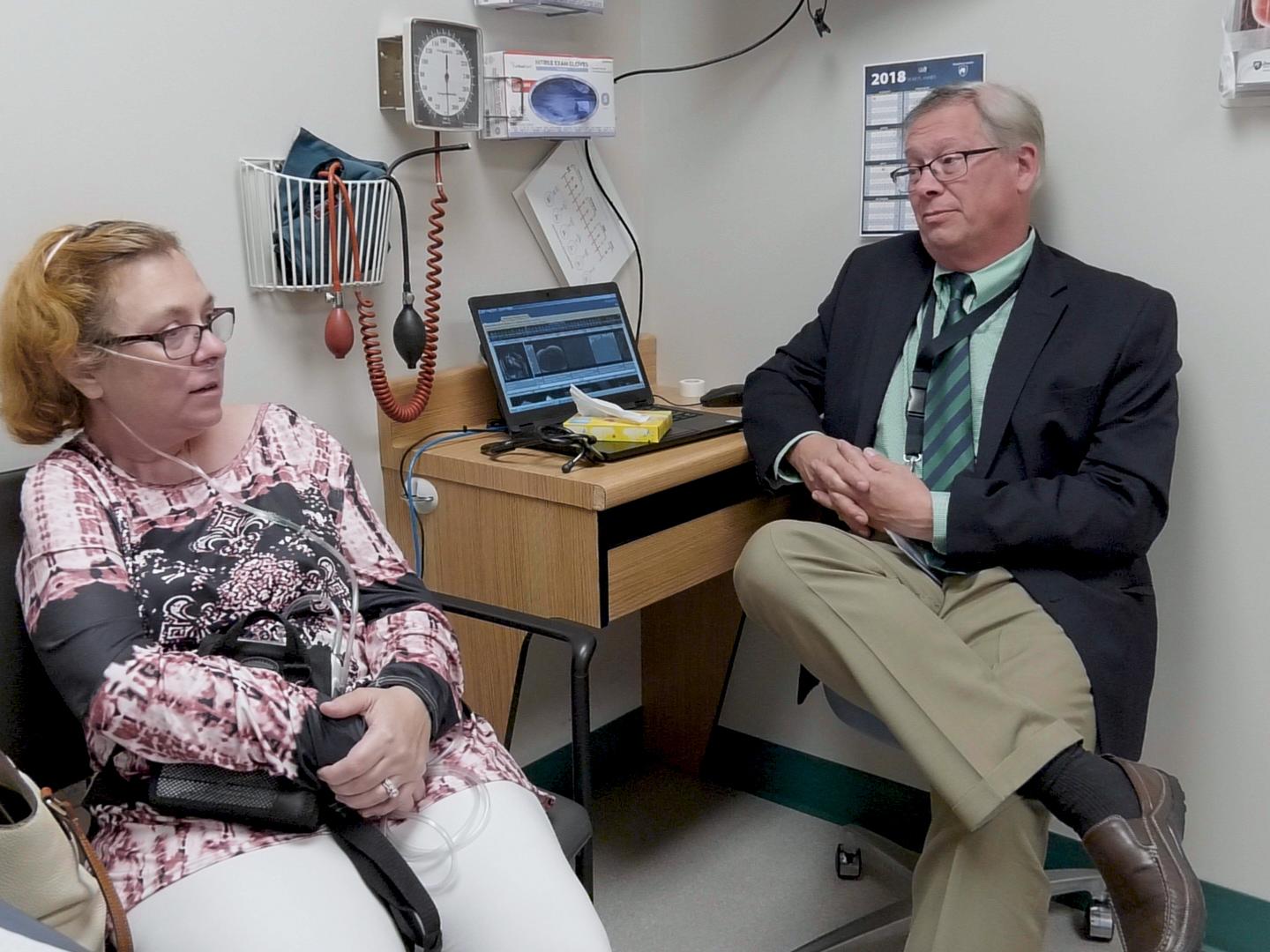 A man and a woman sit in a clinic room, talking. A laptop computer is on a desk and a blood pressure cuff, box of surgical gloves and other clinical items are nearby.