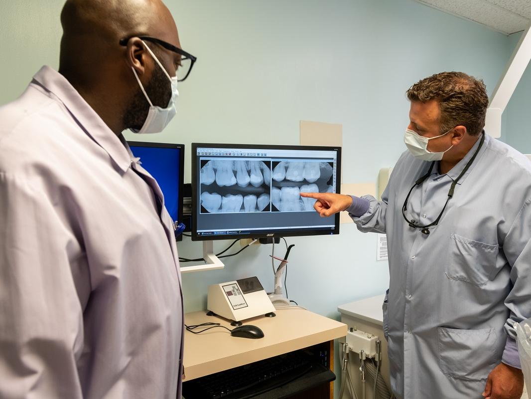 Dr. Larry Trubilla, wearing a lab coat and glasses hanging from his neck, points at the illuminated X-Ray image of a set of teeth. Dental resident Jowhar Brown, wearing glasses and a surgical mask, looks on.