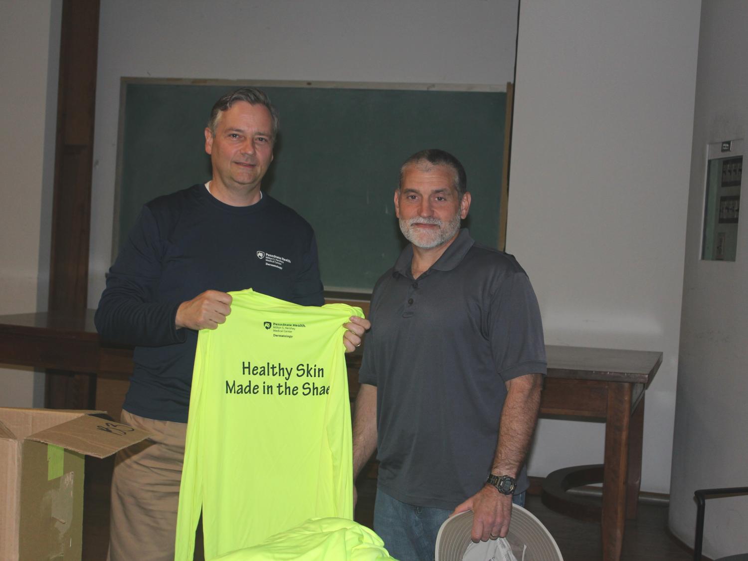 Two men pose for a photo. The man on the left holds a neon yellow shirt with the words “Health Skin Made in the Shade” printed on it. A table and chalkboard are in the background.