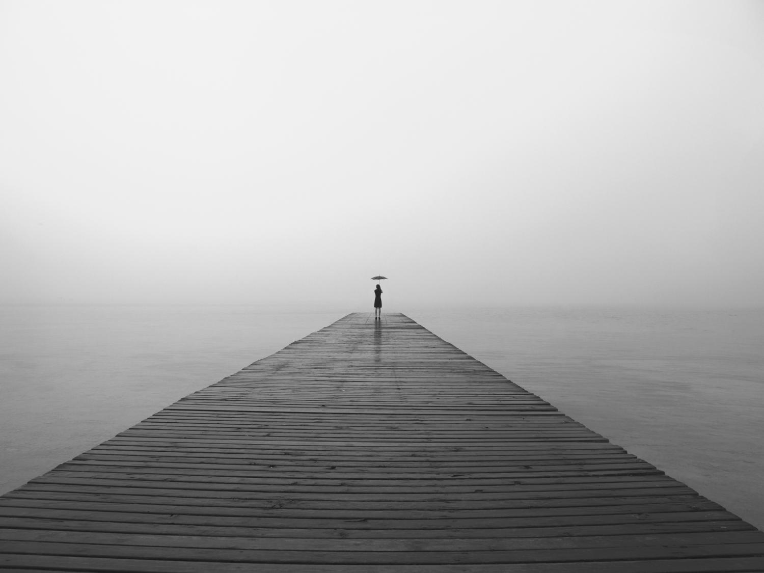 a long peer over water with a very small person at the very end with a black umbrella