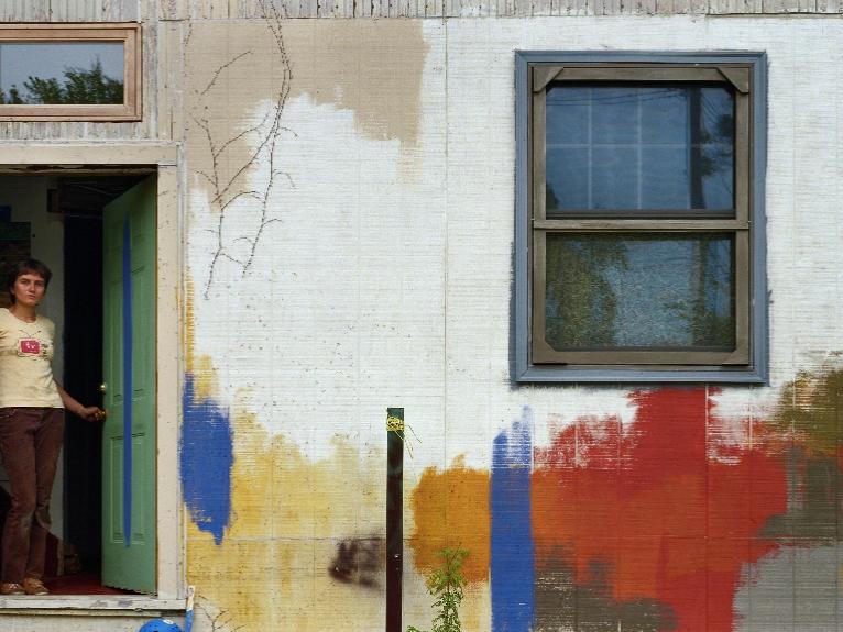 A woman stands in the doorway of a dilapidated house.