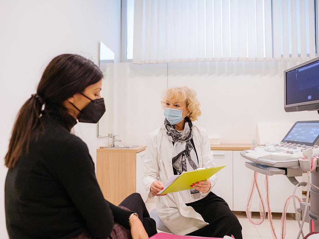 A patient wearing a mask has a conversation with a doctor wearing a mask in an exam room.