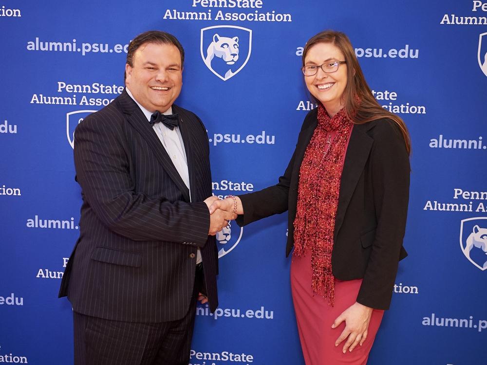 A woman in a dress and blazer shakes hands with a man in a suit with a bow tie. Behind them is a surface covered in the logo and web address for the Penn State Alumni Association.