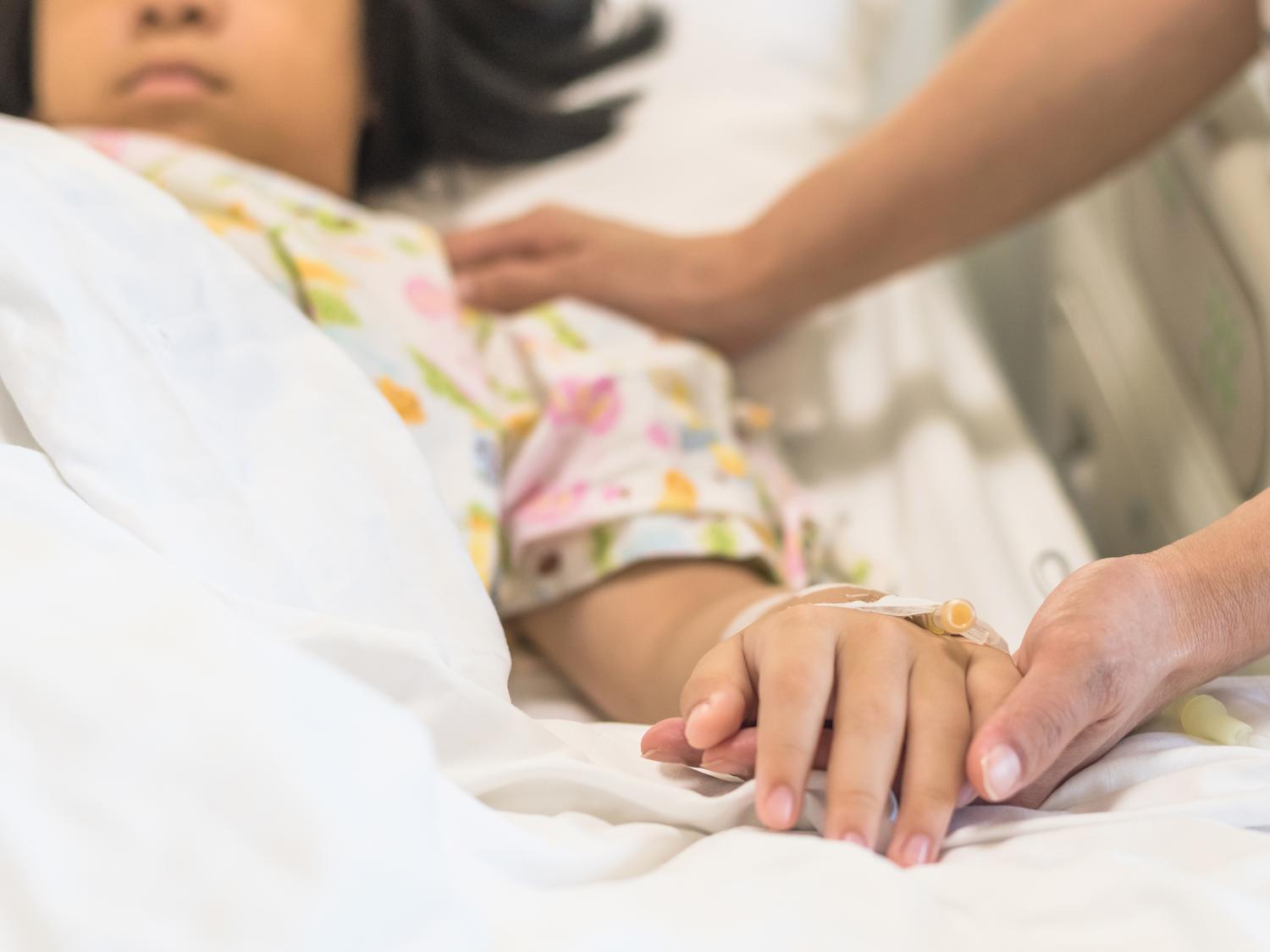 A pediatric cancer patient lays in a hospital bed, while holding hands with a caregiver.