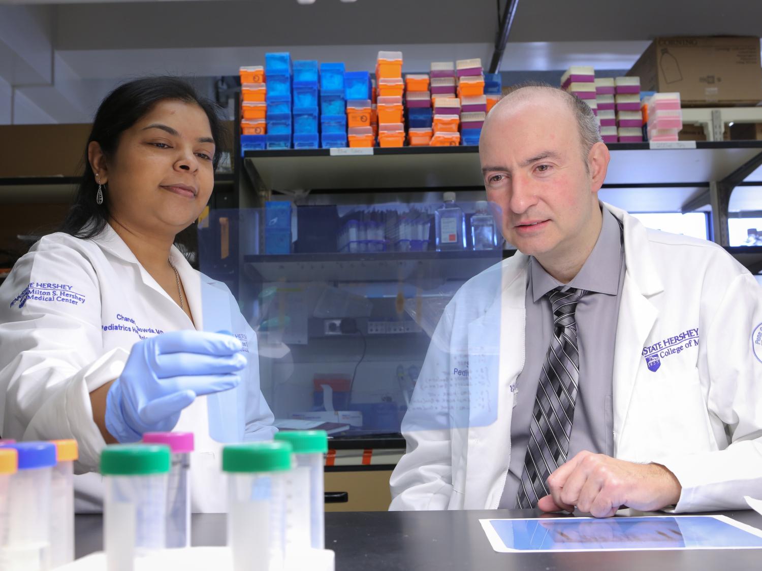Dr. Chandrika Gowda and Dr. Sinisa Dovat are seated at a lab workbench, looking at the camera. Gowda is on the left; Dovat on the right. Both are wearing white lab coats.