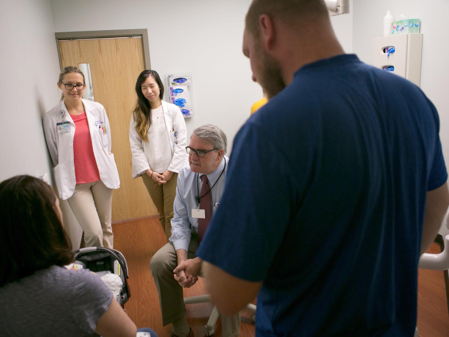 In a clinic room, a man in a tie speaks with a woman who’s holding an infant and facing away from the camera. Three other people are in the room, standing and looking on.