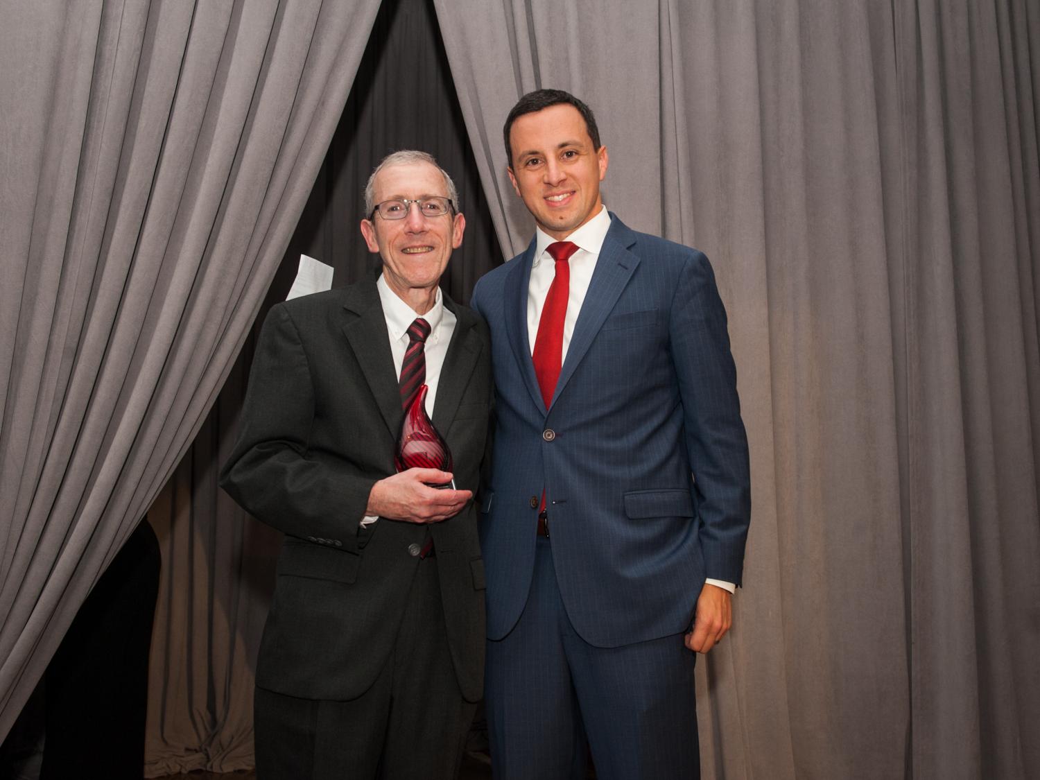 Two men in suits pose for a photo. The man on the left holds a glass trophy fashioned in the shape of a flame. Gray and dark curtains are the backdrop.