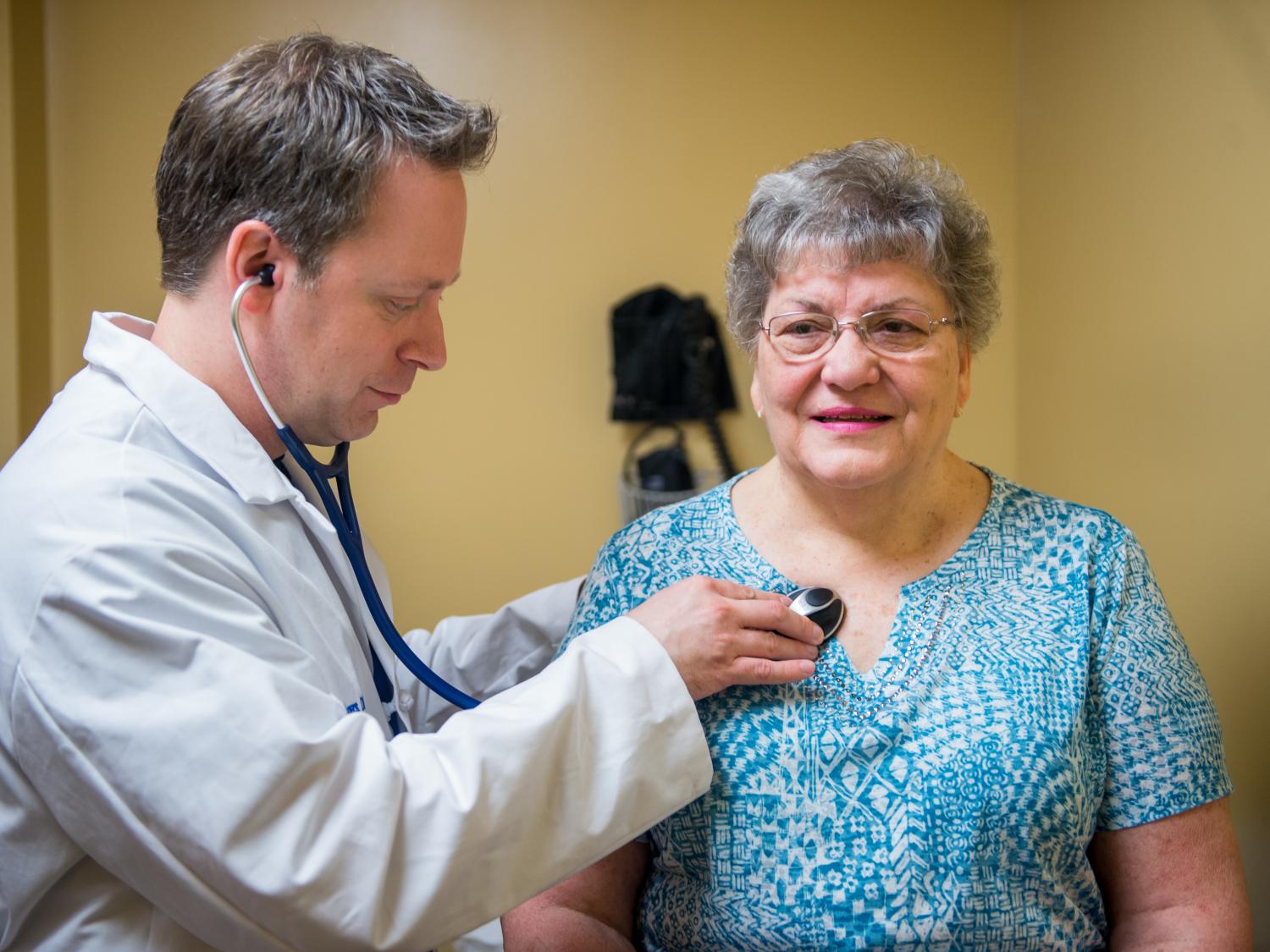 A male physician in a white coat uses a stethoscope to examine a female patient. Small medical equipment is mounted to a wall in the background.