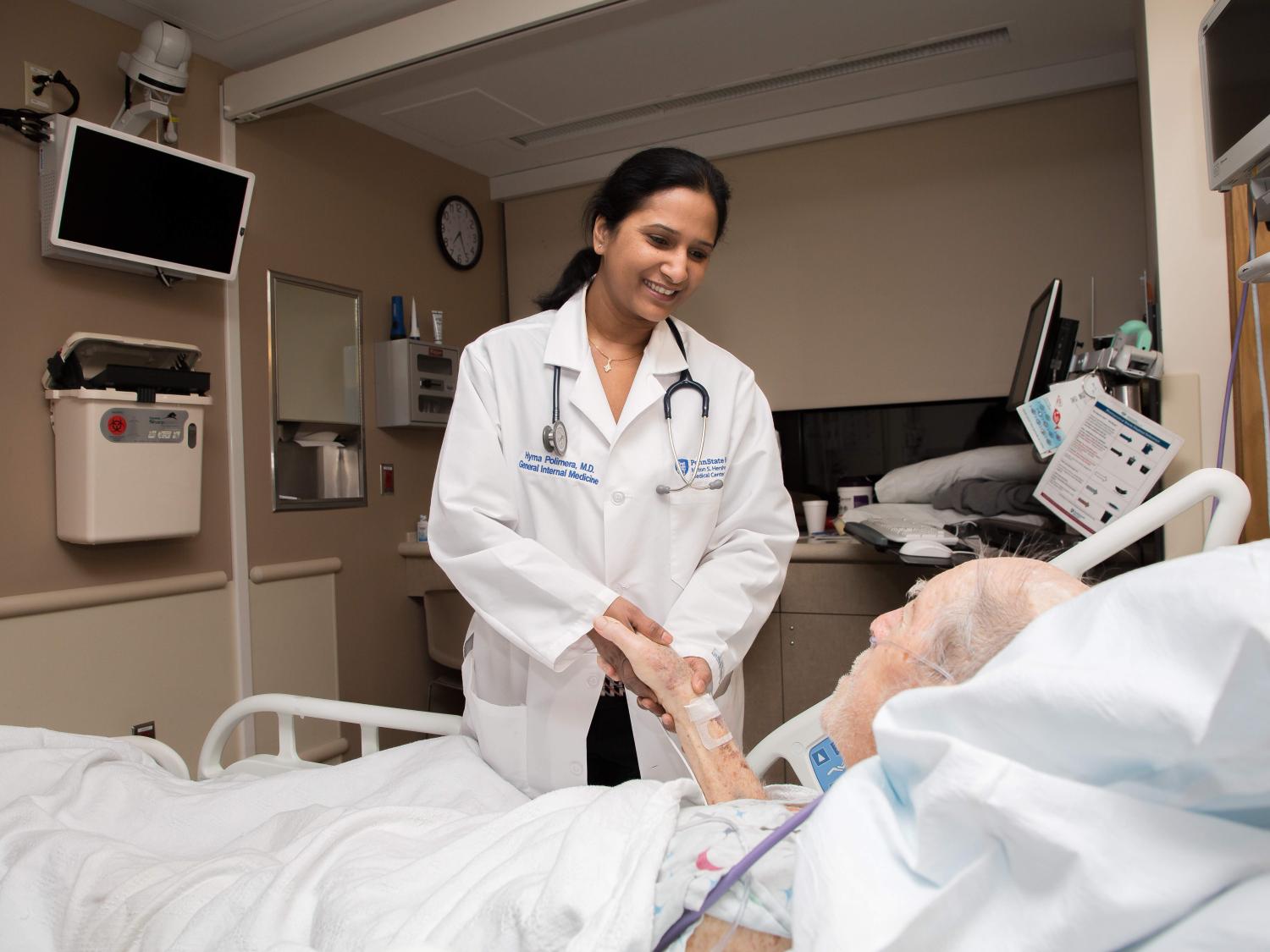 A female physician wearing a white coat and a stethoscope around her neck stands at a patient's bedside, holding his right hand. The patient is reclined in his bed.