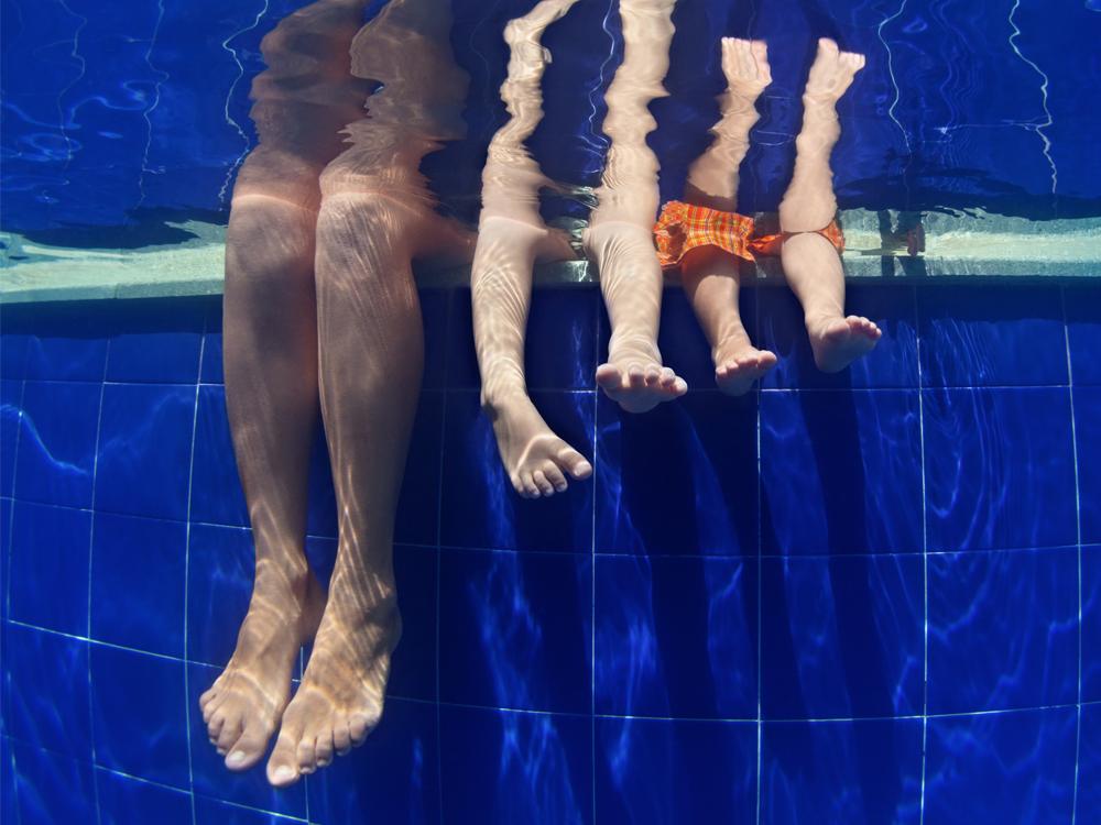 View from underwater of three pairs of legs. All three people are sitting on the edge of the pool. The side of the pool is blue tile.