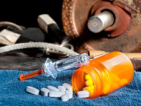 A pill bottle and syringe in the foreground; weights and other sports equipment are in the background.