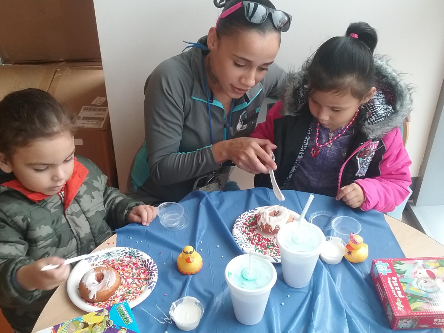 A woman and two children are seated at a small round table, decorating cake donuts using white icing and multi-colored sprinkles.