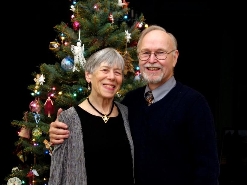 A man and woman stand and smile in front of a Christmas tree.