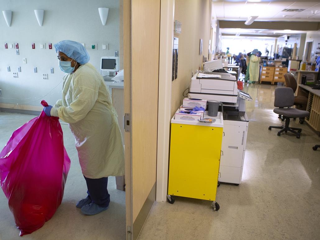 Woman standing in yellow gown, blue hair cover, blue facemask, blue gloves and blue shoe covers holds a red bag while removing red-bag waste from a patient room.