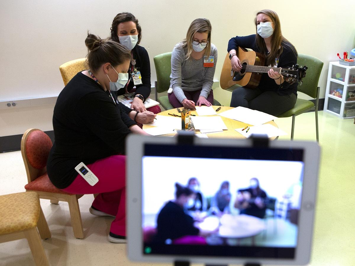 Three Penn State Children’s Hospital staff members sit around a child-sized table, coloring with crayons and paper while music therapist Devon Springer strums a guitar. They are all wearing masks. 