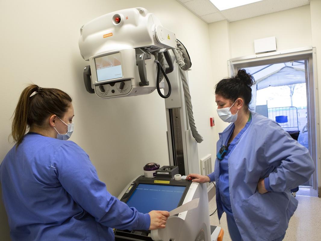 Two radiologic technologists at Hershey Medical Center set up a portable X-ray machine. They are standing by the machine wearing face masks and scrubs. The machine has a large camera on a stand.
