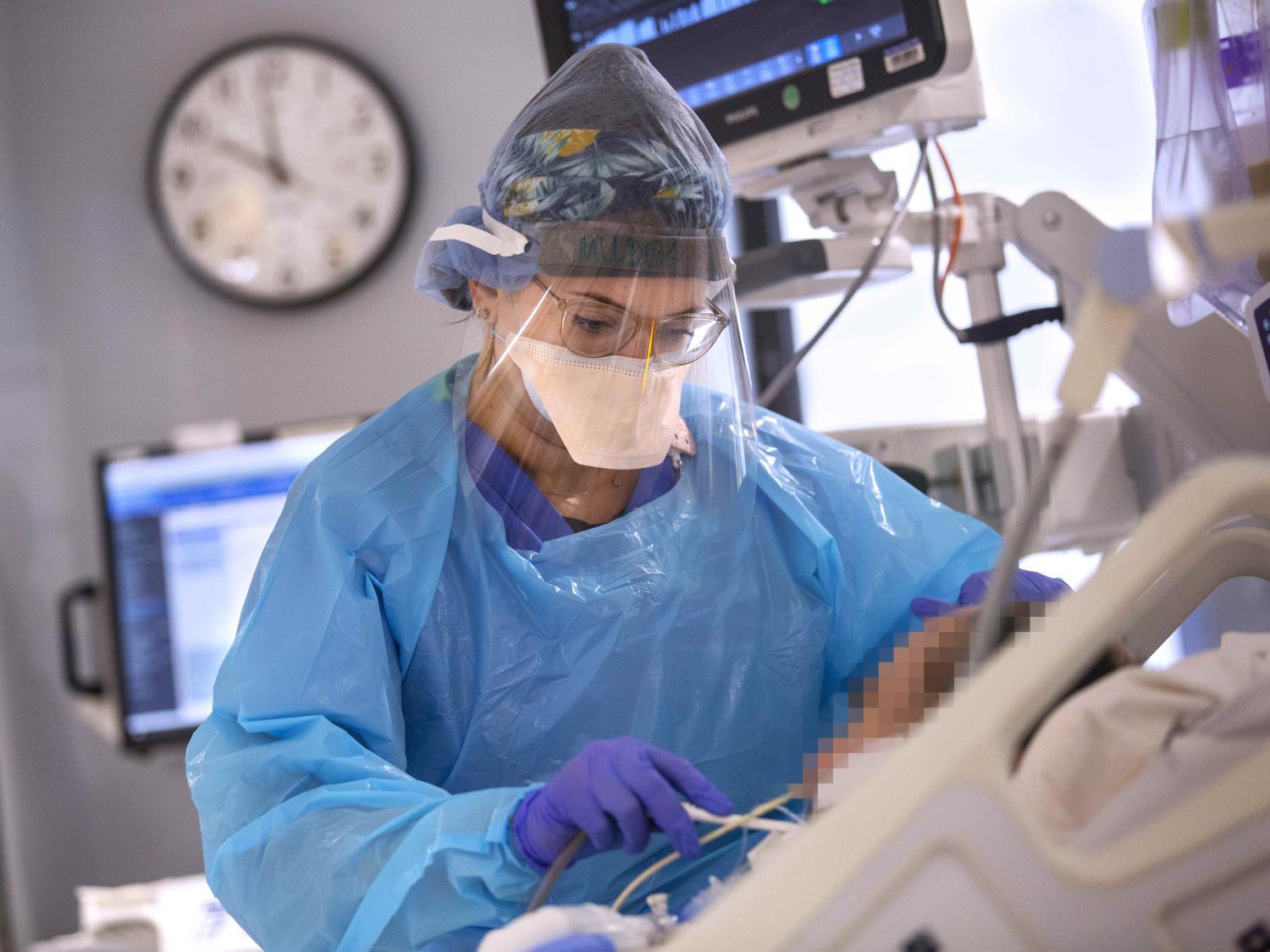 A female nurse in personal protective equipment (PPE) leads over the hospital bed of a patient, holding a toothbrush in her hand and using it to brush the patient's teeth.