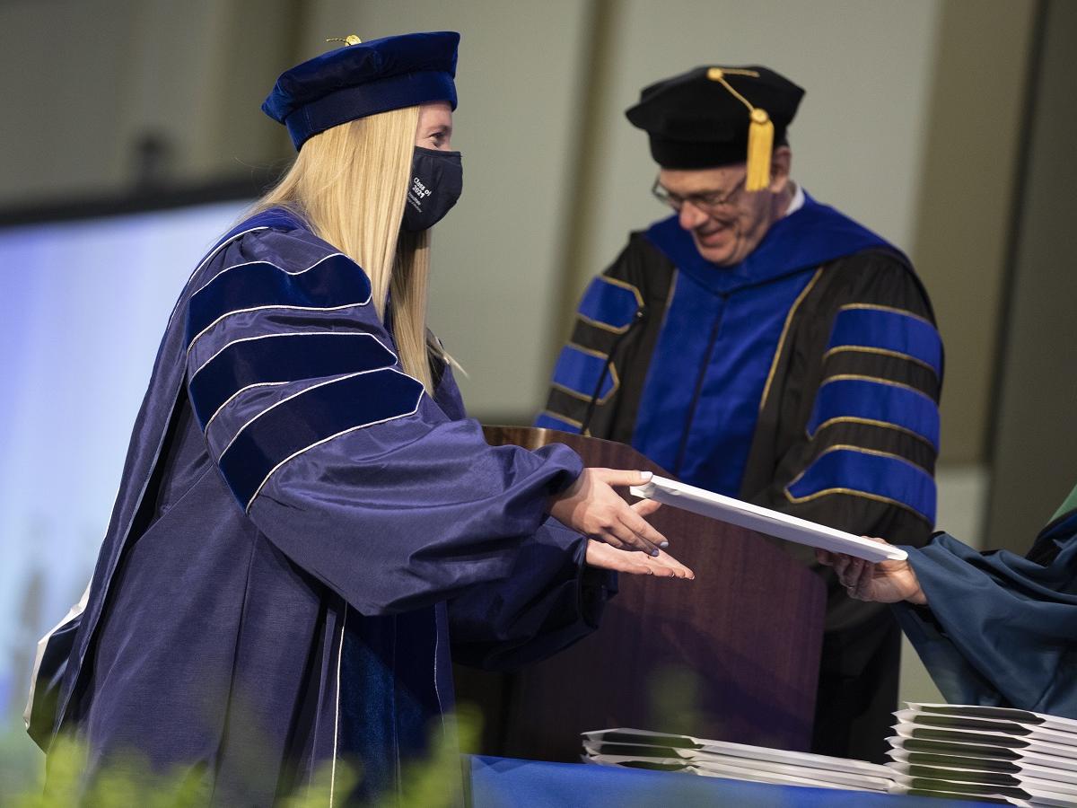 A woman in a graduation cap and gown receives an envelope from someone off camera. Behind her, a man at a lectern in a cap and gown smiles downward.