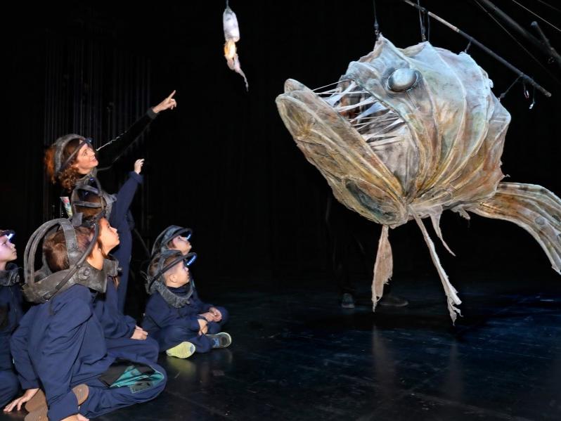 A group of young actors crouch down near a puppet resembling a deep-sea prehistoric fish.