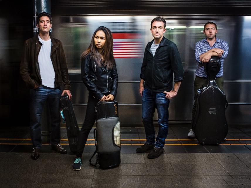 Four musicians dressed in street clothes stand on a subway platform.