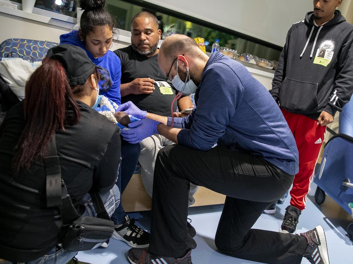 A man and a woman sit on a couch, looking downward at the face of a child in the woman’s lap. In front of them, a doctor in a surgical mask and rubber gloves bows his head as holds the end of a stethoscope to the baby’s chest.