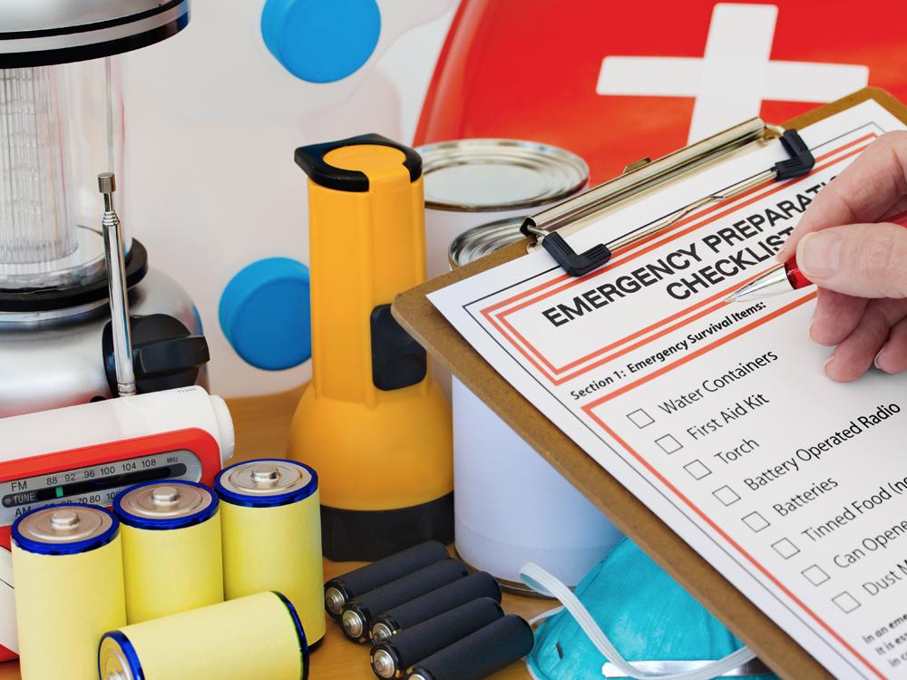A hand holds a pen above a clipboard bearing a sheet reading “Emergency Preparation Checklist.” Items such as batteries, a flashlight and a radio are in the background.