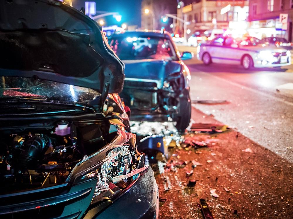 Two cars are parked along the side of a busy city street, each with damage to their front ends from an apparent accident. Bright lights are in the background.