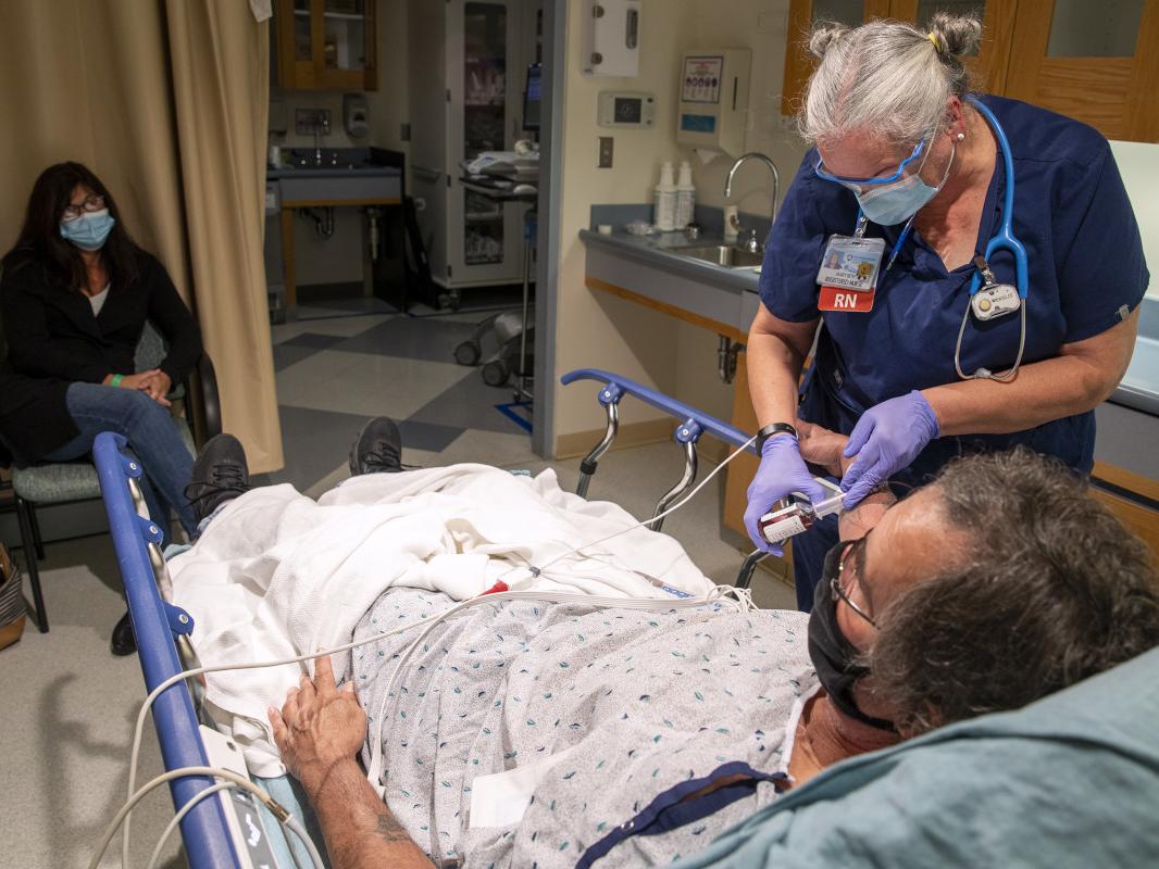 A female nurse administers medication at the bedside of a male patient in an Emergency Department room at Hershey Medical Center. A woman is seated in a chair nearby, looking on.