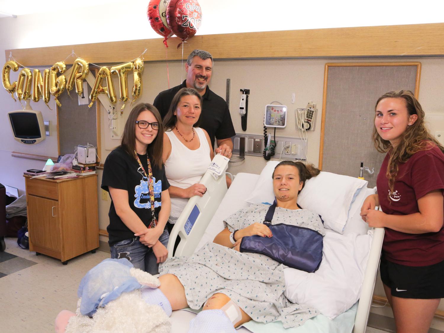 A teenage girl is in a hospital bed, wearing a hospital gown, with her left arm in a blue sling. Three people stand on one side of the bed, one person on the other. Gold balloon letters spelling the word "CONGRATS" are on the wall behind them.