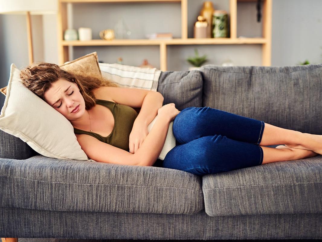 A woman in pain lies on her side on a couch pressing a cushion to her midsection.