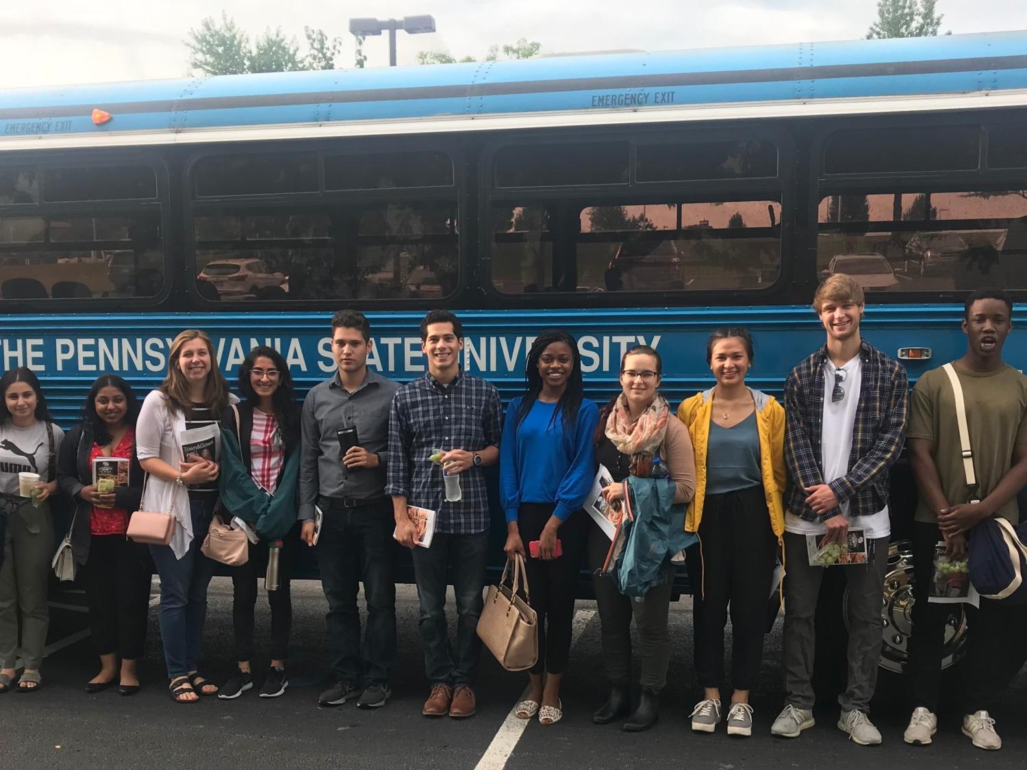 Thirteen people dressed casually stand in a row in front of a bus that has a sign “The Pennsylvania State University” on it. A male professor on the left is wearing a lanyard and name badge.