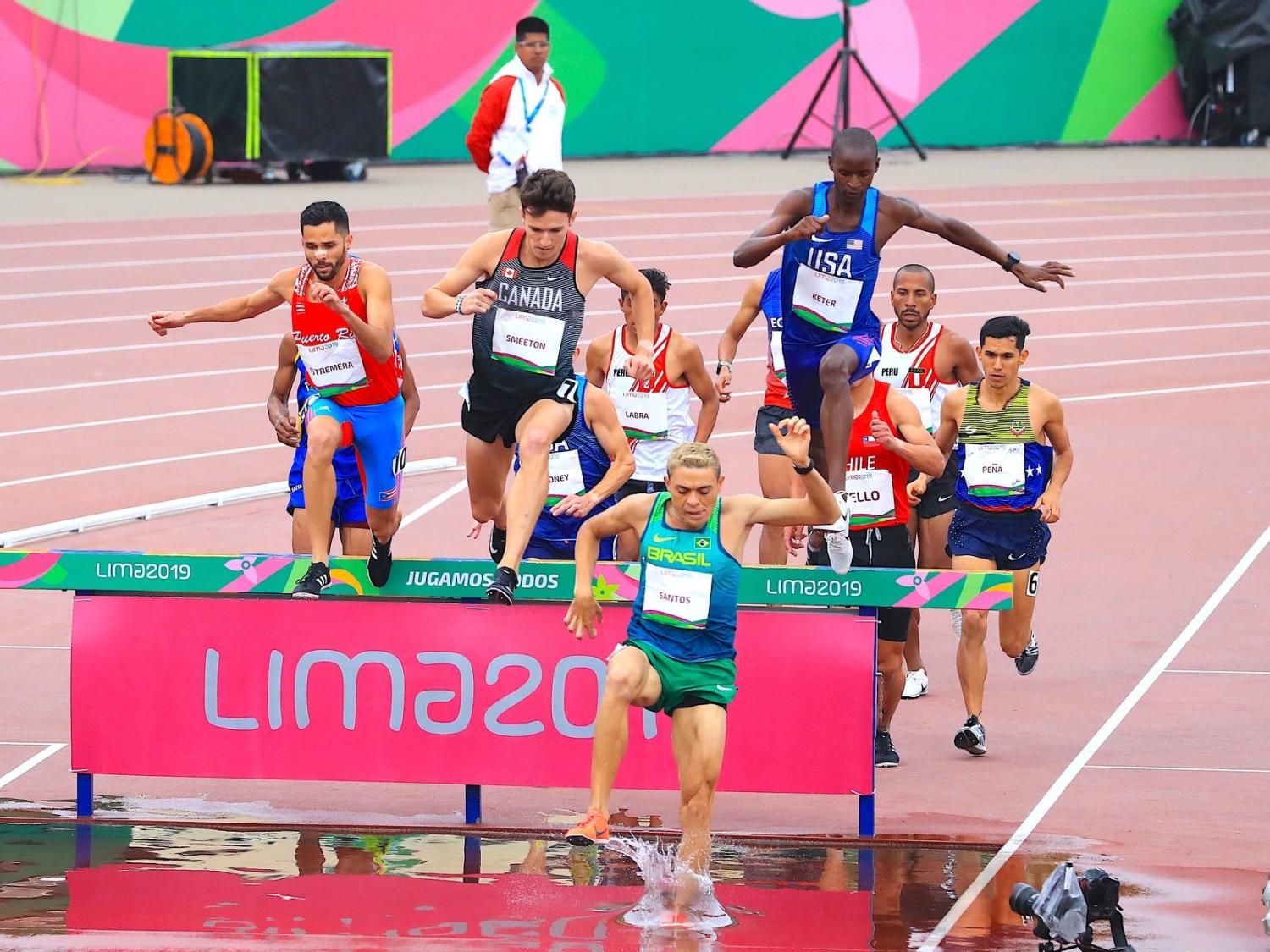 Runners jump over a hurdle and into water during a steeplechase race.