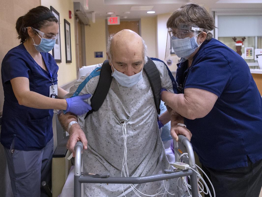 A man in a hospital gown rises out of a wheelchair to a standing position, leaning on a walker. Several wires emerge from his gown. Two staff members, one on each side, are bracing the man’s upper arms.